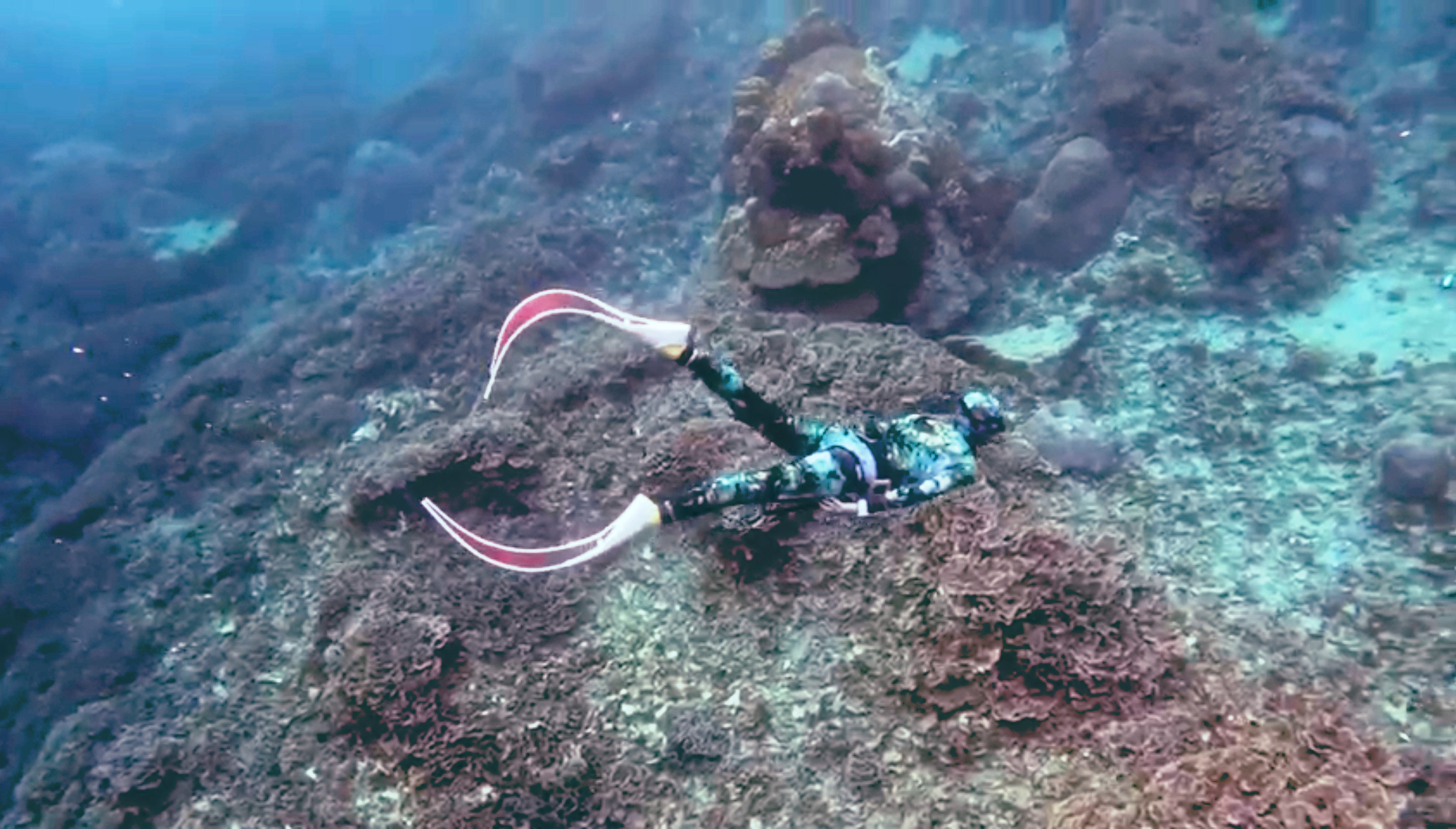 Freediver in a camouflage wetsuit gliding over a coral reef with long fins